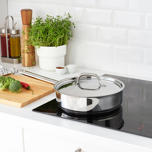 Cocina moderna con sartén de acero inoxidable en estufa de inducción, tabla de cortar con verduras, condimentos y una planta.