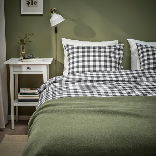 Acogedora cama con edredón EKTANDVINGE de cuadros blancos/negros, manta verde, almohadas blancas. La mesita de noche sostiene libros, lámpara, planta.