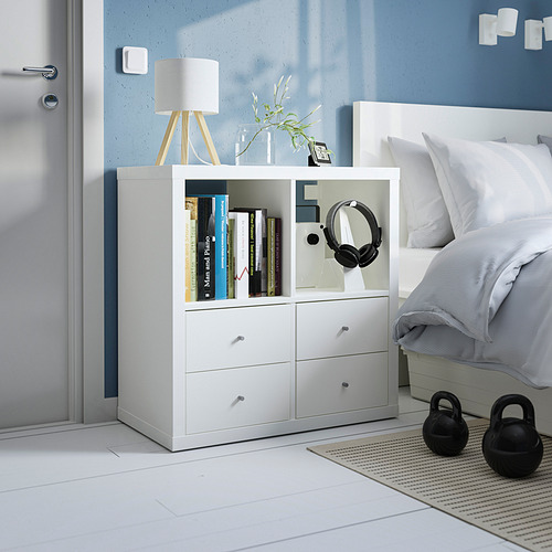 Unidad de almacenamiento KALLAX blanca al lado de la cama, con libros, auriculares y lámpara. Más pequeño que la cama, cabe perfectamente en la esquina.