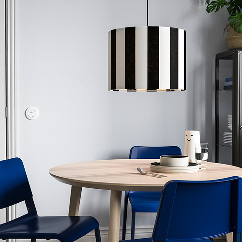 Comedor minimalista con mesa de madera clara, sillas azules y una lámpara colgante única a rayas blancas y negras, más grande que la planta cercana.