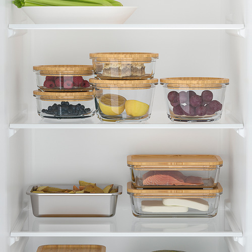 Estante de la nevera con recipientes de vidrio apilados para alimentos, frutas y una lata para preparar comidas, que muestra el almacenamiento organizado de alimentos.