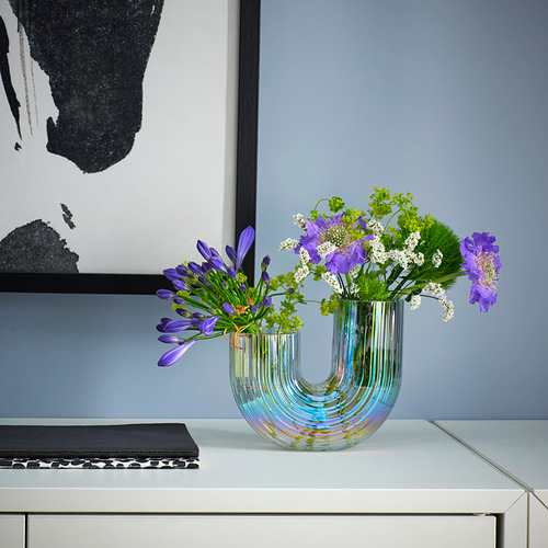 Un jarrón RÄFFELBJÖRK con flores moradas en forma de arco iris, sobre una mesa blanca junto a un libro y un cuadro.