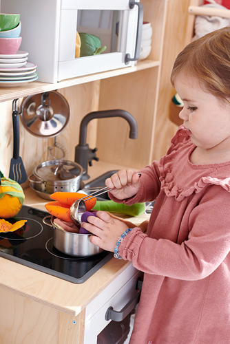 Una niña juega con la cocina de juguete DUKTIG, cortando verduras de color naranja en una estufa negra, vestida con un suéter rosa.
