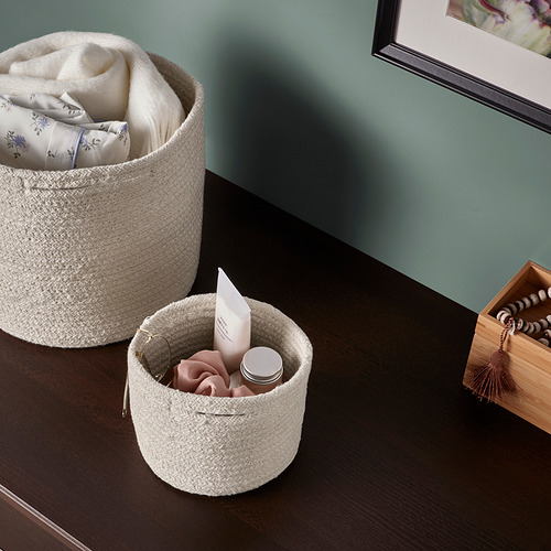 Dos cestas de almacenamiento de algodón tejido sobre una mesa de madera, una contiene toallas, la otra artículos pequeños y cosméticos.