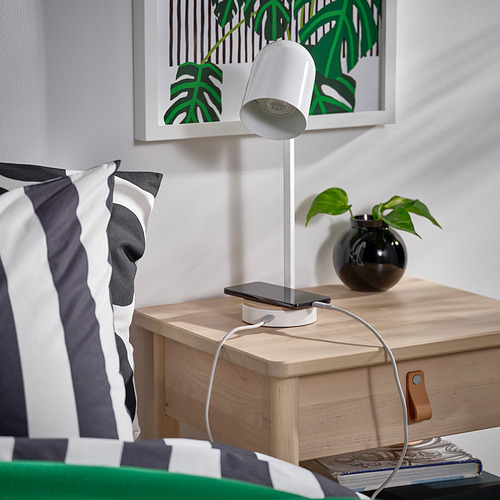 Una lámpara de escritorio blanca con base de madera y puerto de carga USB-C se encuentra junto a una cama, cargando un teléfono inteligente.