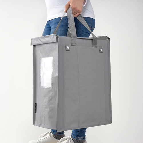 Persona sosteniendo una bolsa DIMPA gris por asas. La bolsa tiene un bolsillo transparente para etiquetar, adecuado para la clasificación de residuos.