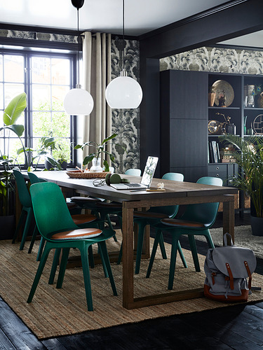 Comedor moderno con mesa de madera, sillas verdes y dos lámparas colgantes de globo blanco. Paredes oscuras, cortinas estampadas y plantas.