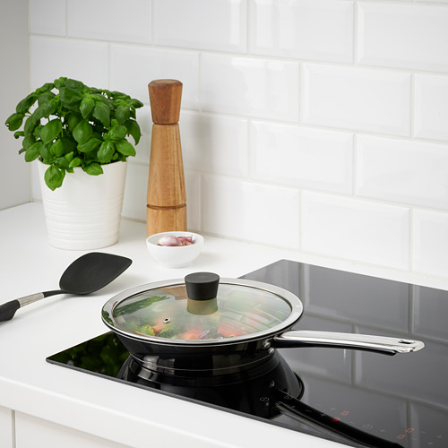 Escena de cocina: sartén en estufa con tapa de vidrio, planta de albahaca y molinillo de pimienta de madera.