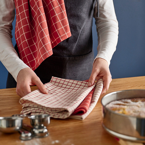 Una persona dobla paños de cocina a cuadros rojos y blancos sobre una mesa de madera, usándolos para secar platos y limpiar superficies.