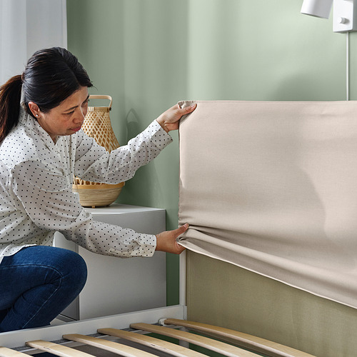 Una mujer ajusta el cabecero de una cama KLEPPSTAD. La cama tiene una moderna estructura de metal y un acogedor cabecero tapizado.