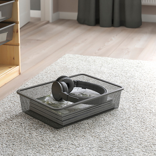 Caja de almacenamiento de malla sobre alfombra, que contiene auriculares y caja. Forma parte de la serie TROFAST para la organización de artículos.