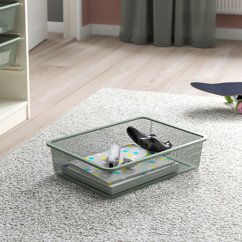 Cesta de almacenamiento de malla sobre alfombra, que contiene accesorios remotos y electrónicos, parte de la serie TROFAST para organizar artículos.