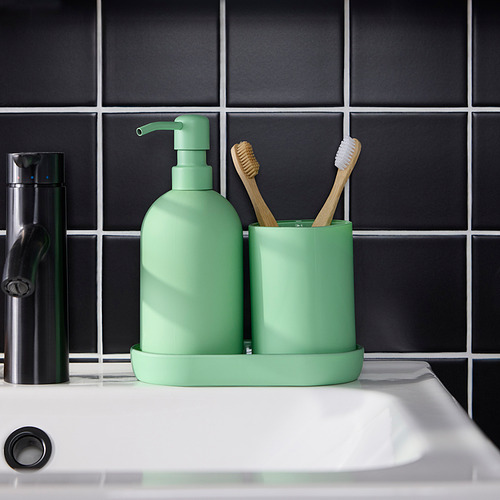 Dispensador de jabón verde y soporte para cepillos de dientes en fregadero blanco. La bandeja mantiene los artículos organizados. Contra una pared de azulejos oscuros.