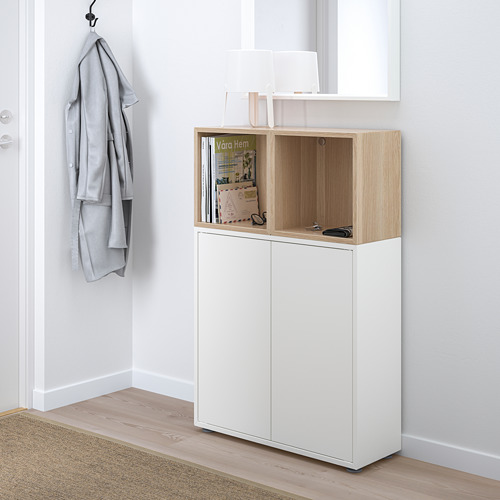 Mueble de almacenaje EKET blanco con encimera de madera, secciones abiertas/cerradas, exhibiendo una revista y tijeras, suéter gris en gancho.