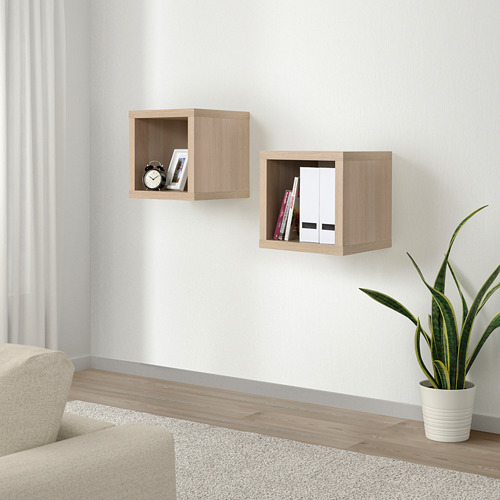 Estanterías de pared KALLAX, madera clara. Pequeños cubos cuadrados sostienen libros y relojes. Al lado de una silla y una planta de color beige.