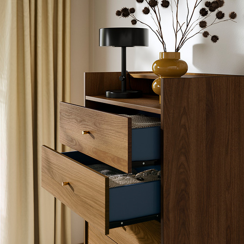 Cómoda de madera con cajones azules, cajón abierto, lámpara negra, jarrón amarillo, cortinas beige.
