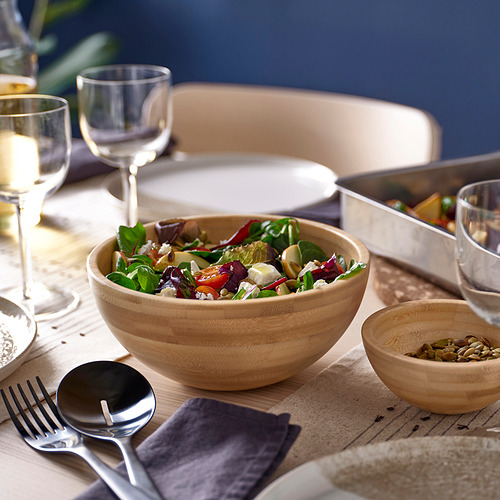 Gran cuenco de madera con ensalada en la mesa. Cerca de copas de vino, platos y un tazón más pequeño con semillas. Acompañado de utensilios para servir y servilletas.