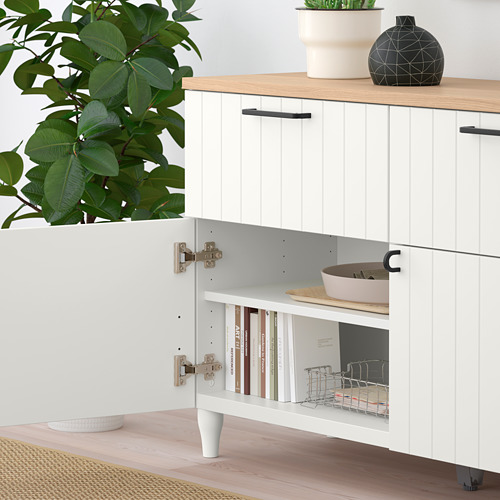Moderno mueble de almacenaje con estantería abierta que contiene libros y cesta, armario cerrado encima y artículos decorativos en la parte superior. En las inmediaciones hay plantas y un cuenco.