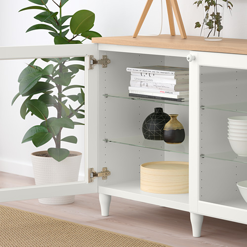 Armario blanco con puerta de cristal, donde se exhiben libros, jarrones y cuencos. Planta de interior a su lado.