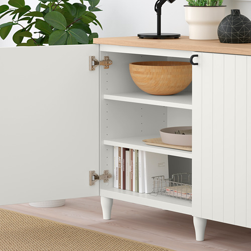 Aparador blanco BESTÅ con puerta abierta que muestra un cuenco de madera, libros y organizador de platos en estantes. Tapa decorada con plantas y lámpara.