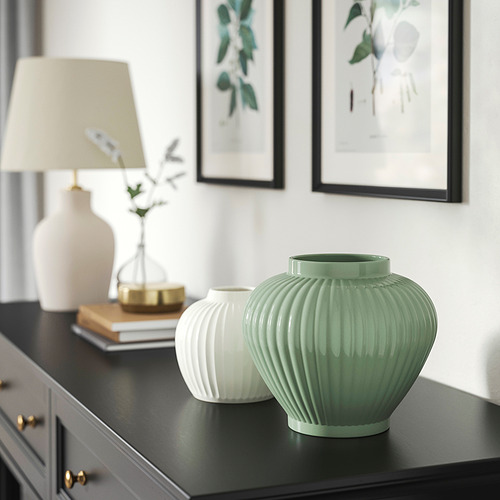 Jarrones PELARBJÖRK de color verde grisáceo en estantería negra con libros, lámpara y decoración dorada. El jarrón verde más grande contrasta con uno blanco más pequeño.