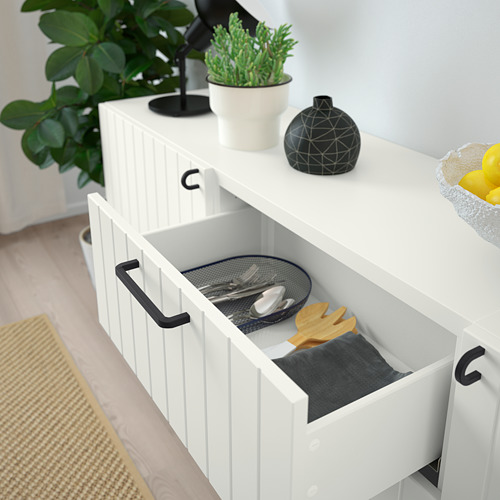 Aparador blanco BESTÅ con cajones abiertos, que exhibe utensilios de cocina, colocado contra una pared con plantas decorativas y limones.