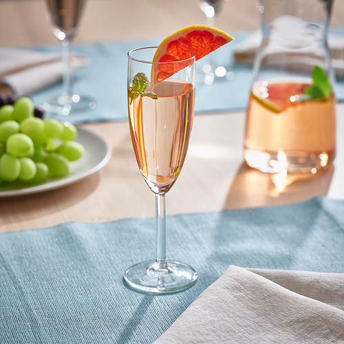 Un vaso alto y transparente con un borde inclinado sostiene una bebida burbujeante adornada con una rodaja de pomelo, colocada sobre un mantel azul claro cerca de uvas verdes.