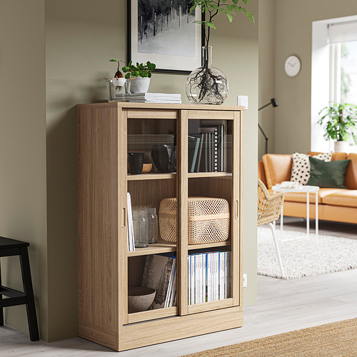 Librería TONSTAD de roble con puertas de cristal en acogedor salón, decorada con plantas y jarrones.