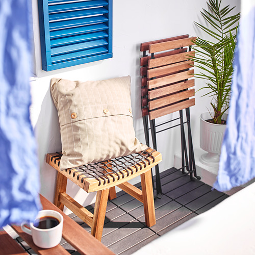 Escena de balcón con taburete de madera STACKHOLMEN, cojín beige, taza de café, planta en maceta, persianas azules y cortinas azul claro.