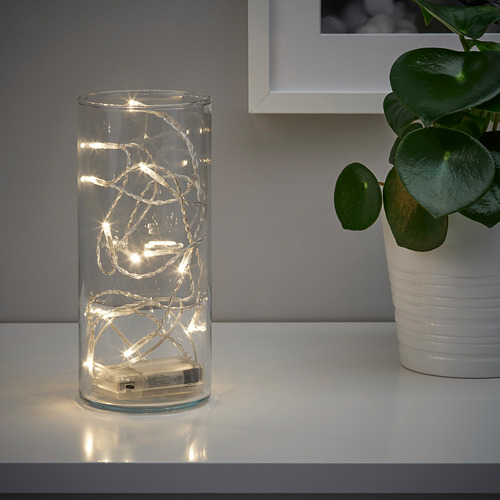 Un jarrón transparente contiene luces LED blancas cálidas, creando un ambiente acogedor. Una planta en maceta se encuentra a su lado sobre una superficie blanca.