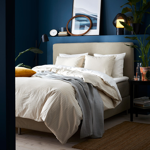 Dormitorio moderno con paredes de color azul intenso, con una cama tapizada en color beige con ropa de cama de rayas blancas. La decoración incluye plantas, lámparas y arte mural.