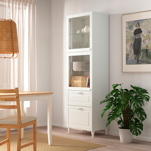 Gabinete blanco con puerta de vidrio BESTÅ en esquina, exhibiendo platos, cerca de la mesa de comedor y la planta.