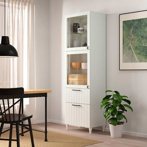 Armario alto blanco BESTÅ con puertas y estantes de vidrio, con artículos decorativos, junto a mesa y sillas negras, planta verde.