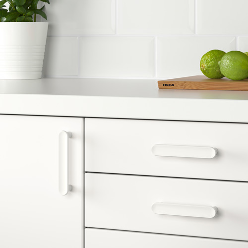 Mueble de cocina blanco con tres cajones, pomos sencillos, planta verde, limas y tabla de cortar sobre encimera.