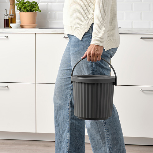 Persona que sostiene un contenedor gris con asa, ideal para residuos pequeños como metal o baterías. El cubo tiene una tapa extraíble para ocultar los residuos.