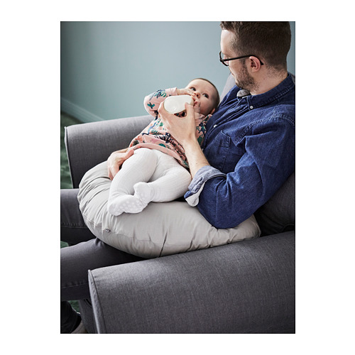Un hombre sostiene a un bebé con una almohada de lactancia en un sofá gris. La almohada es blanca y amortigua al bebé para alimentarlo.