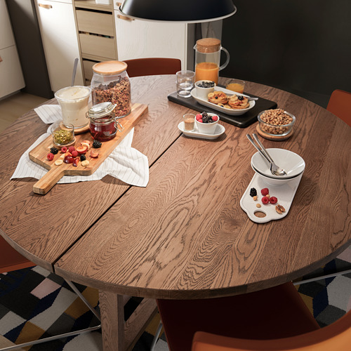 Mesa de comedor redonda de madera con chapa de roble, rodeada de sillas de color naranja, decorada con productos para el desayuno como fruta, yogur, granola y jugo.