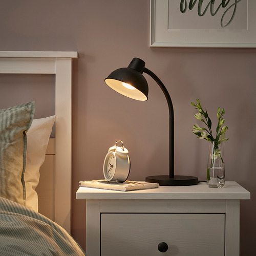 Dormitorio moderno con lámpara SKURUP en mesita de noche, libros y plantas iluminados, almohada a rayas en la cama, pared marrón cálido.