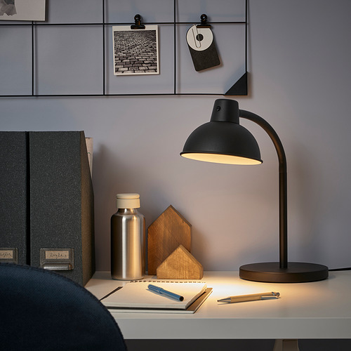 Montaje de escritorio minimalista con una pequeña lámpara SKURUP negra, que emite una luz brillante, rodeada de libros y decoración.