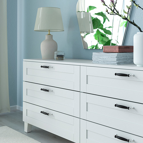 Tocador blanco con detalles oscuros, seis cajones, lámpara, decoración y planta de interior contra la pared azul.