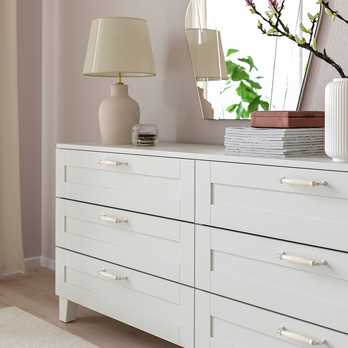 Cómoda blanca con seis cajones, lámpara de luz, libros y jarrón decorativo.