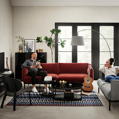 Salón: el hombre lee, la mujer escucha música. Sofás rojos y grises, mesa de centro, plantas, guitarra.