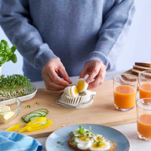 Persona que prepara rodajas de huevo hervido con una práctica cortadora de huevos en una tabla de madera con varios artículos para el desayuno.