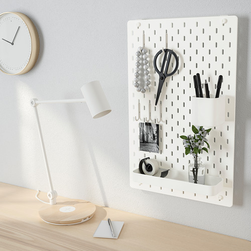 Un organizador de tablero de clavijas con tijeras, bolígrafos, una planta y un espejo, junto a un escritorio con una lámpara y un reloj en la pared.