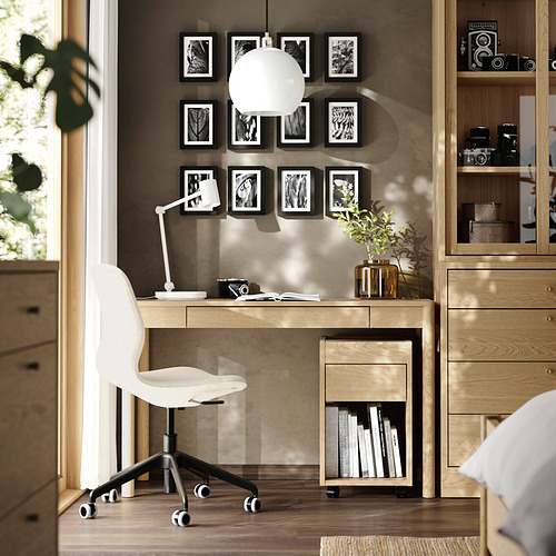 Moderna oficina en casa con escritorio de roble TONSTAD, silla blanca, fotos y lámpara de globo. La luz natural llena la habitación.
