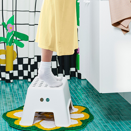 Niño pisando un pequeño taburete blanco BOLMEN, vestido amarillo, alfombra verde, decoración colorida.