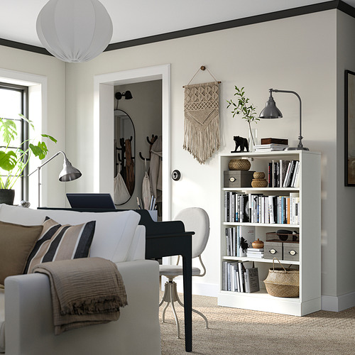 Sala de estar luminosa con decoración moderna: sofá blanco, silla gris, estantería de madera con libros, plantas y una lámpara negra.