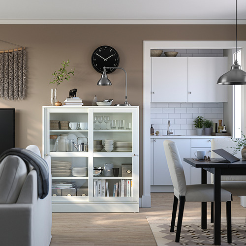 Mueble de almacenamiento HAVSTA blanco con puertas de vidrio, madera de pino y vidrio templado, exhibiendo platos y libros, bajo un reloj negro.