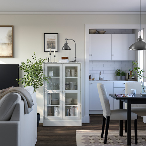 Moderno living comedor con mueble de pino blanco, puertas de cristal. Cocina blanca en forma de L, lámpara gris, mesa de comedor negra con sillas blancas.