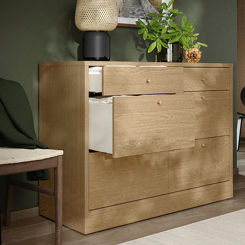 Cómoda de madera natural con cajones abiertos, pared verde, plantas, lámpara en la parte superior.